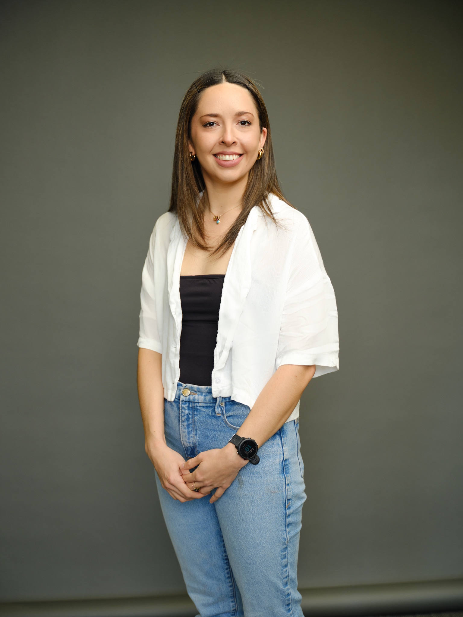 Picture of woman smiling, while wearing a white sweater, brown top and blue jeans. Her hands are clasped in front.  