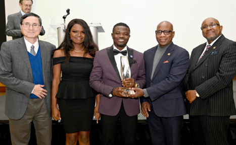 The image shows a group of five people standing together, posing for a photo. The man in the center is holding a large, clear trophy, indicating an award ceremony or recognition event. They are all dressed in formal attire, including suits and a black dress. The setting appears to be indoors, with a podium and a white background, suggesting a formal presentation or event. The group is smiling, indicating a celebratory atmosphere.