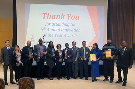 The image shows a group of people standing in a line, posing for a photo on a stage. They are holding awards and certificates, indicating that this is an award ceremony. Behind them is a large screen with a message thanking attendees for participating in the fifth annual Innovation of the Year Awards. The individuals are dressed in formal attire, including suits and dresses, creating a professional and celebratory atmosphere.