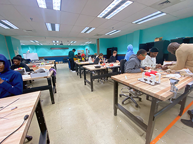 The image shows a classroom or workshop setting with several students sitting at workbenches, engaged in hands-on activities. The room has turquoise walls, fluorescent lighting, and large wooden tables. A teacher is assisting a group of students on the right side of the room. The environment appears collaborative and focused on practical learning.