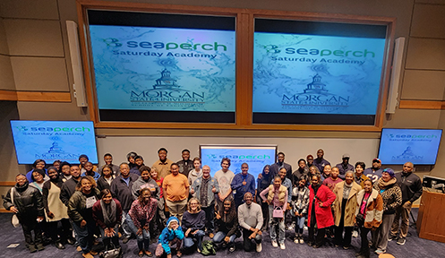 The image shows a large group of people posing for a photo in a lecture hall or auditorium. They are standing in front of a stage with two large screens displaying the logo for SeaPerch Saturday Academy and Morgan State University. The group consists of men, women, and children, suggesting a diverse mix of participants, possibly students, faculty, and community members. The atmosphere appears to be educational and community-oriented.